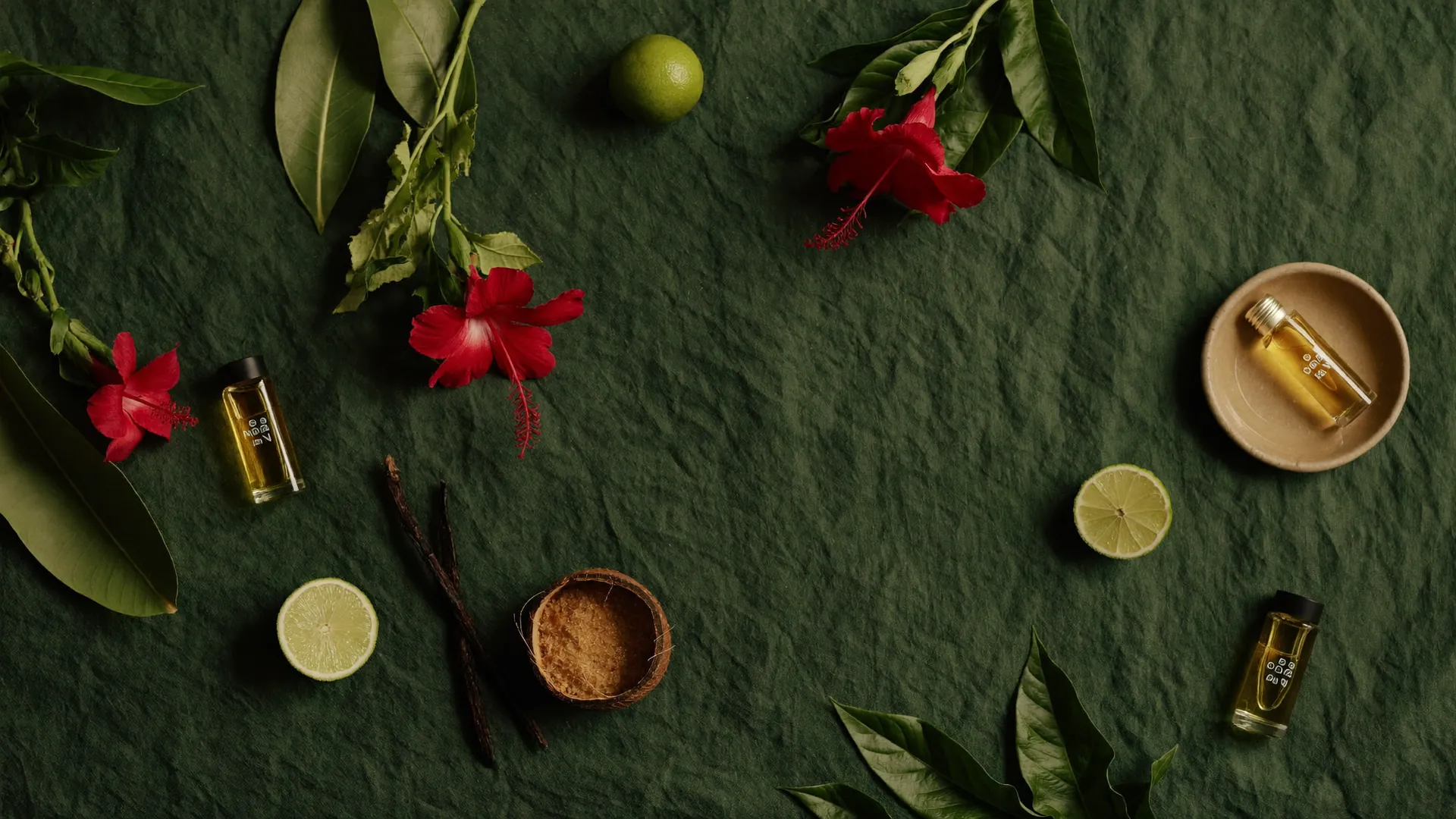 Island botanicals on forest-green linen — hibiscus, lime, vanilla pods, brown sugar, coconut husk and vials of golden oil